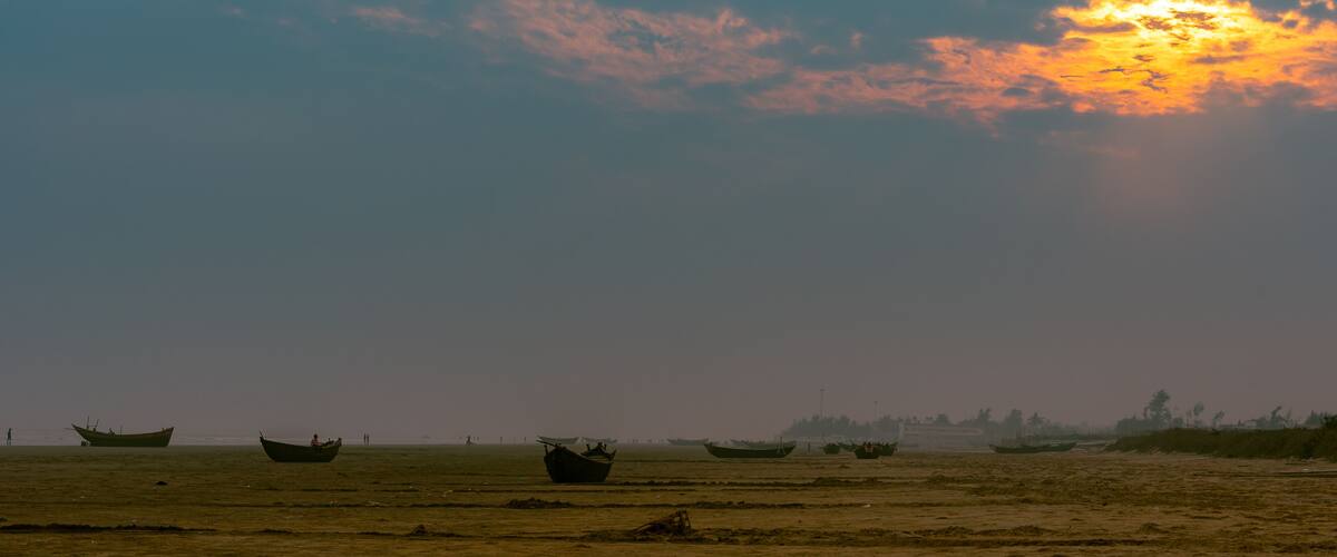 A coastal landscape with several boats anchored on a sandy beach, under a sunset sky.