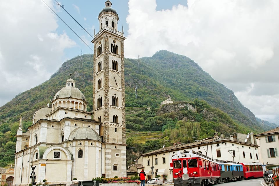 A historic church with a bell tower, a red train, and a mountainous backdrop.