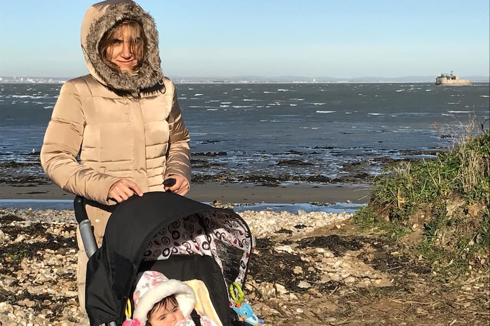 Une personne portant une veste doublée de fourrure, assise au bord d’une côte rocheuse, à côté d’une poussette dans laquelle se trouve un bébé.
