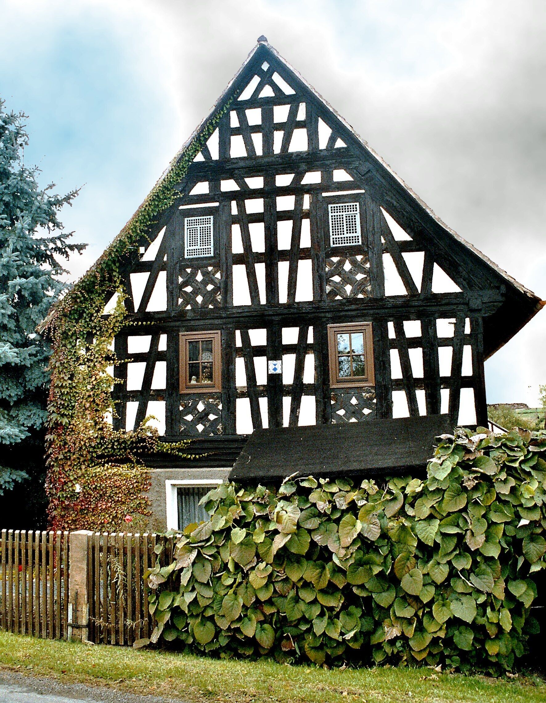 A half-timbered house with a steeply pitched roof, surrounded by a wooden fence and a hedge.
