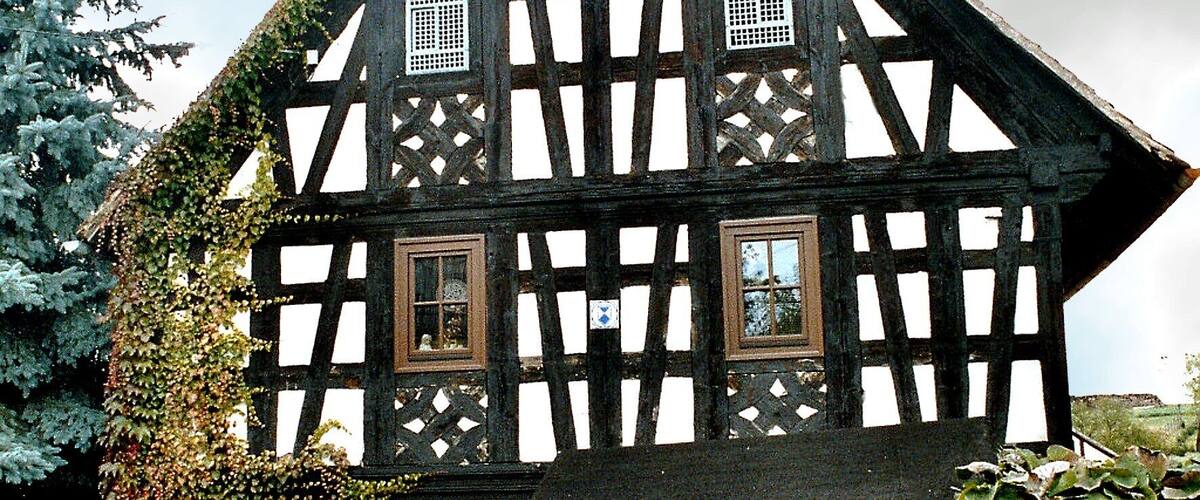 A half-timbered house with a steeply pitched roof, surrounded by a wooden fence and a hedge.