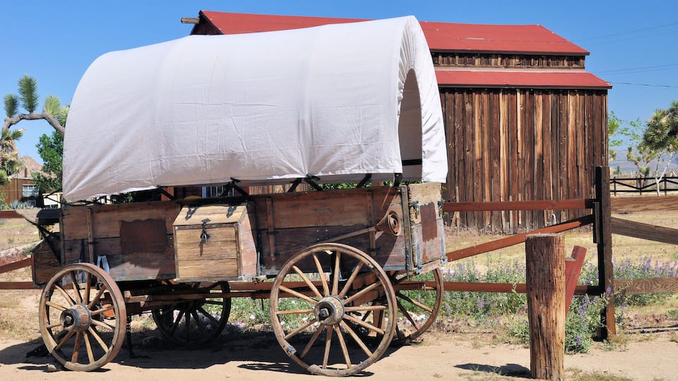 A covered wooden wagon with large wheels parked in front of a wooden building with a red roof.
