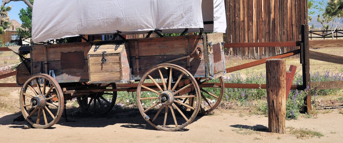 A covered wooden wagon with large wheels parked in front of a wooden building with a red roof.