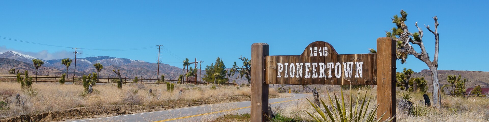 Un letrero de madera con la inscripción '1945 PIONEERTOWN', rodeado de vegetación desértica y un cielo azul despejado.