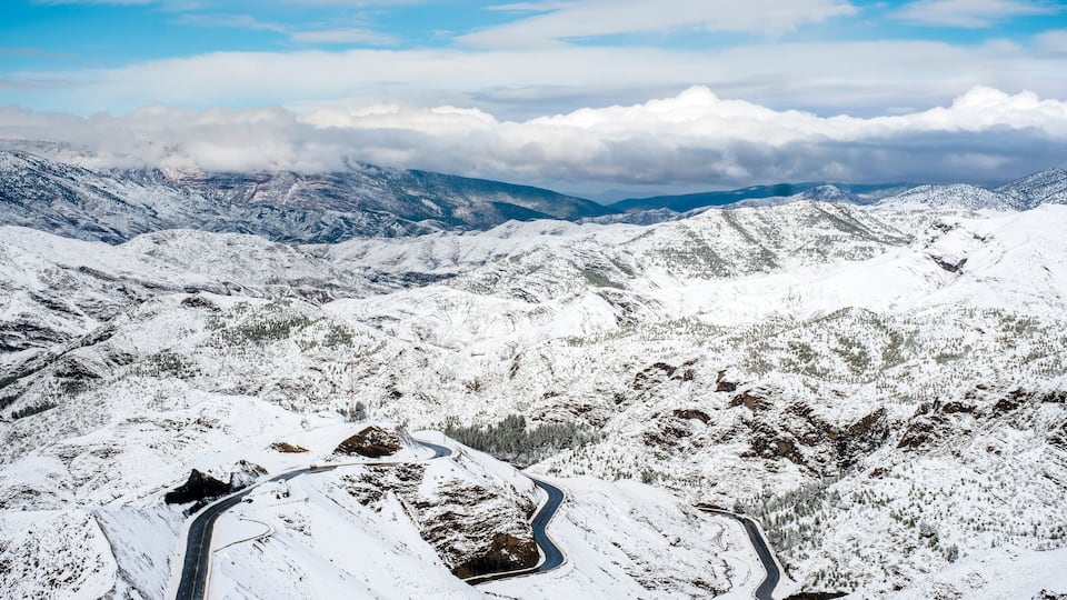 A snow-covered mountain landscape with winding roads and distant clouds.