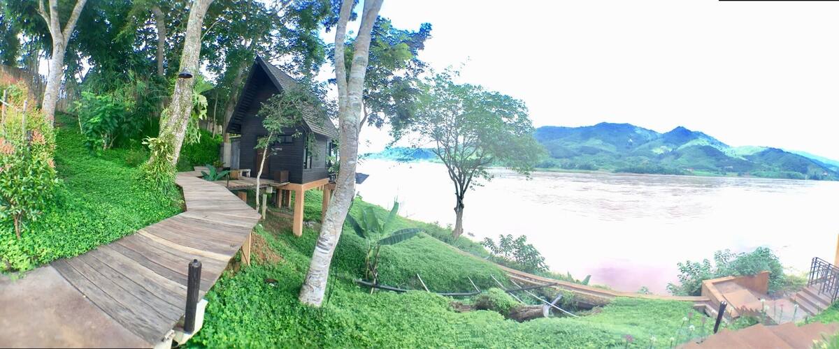 A wooden deck leading to a cabin by a river, surrounded by lush greenery and hills.