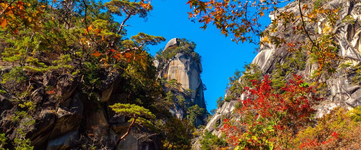A mountainous landscape with a prominent rock formation, dense foliage, and a clear blue sky.