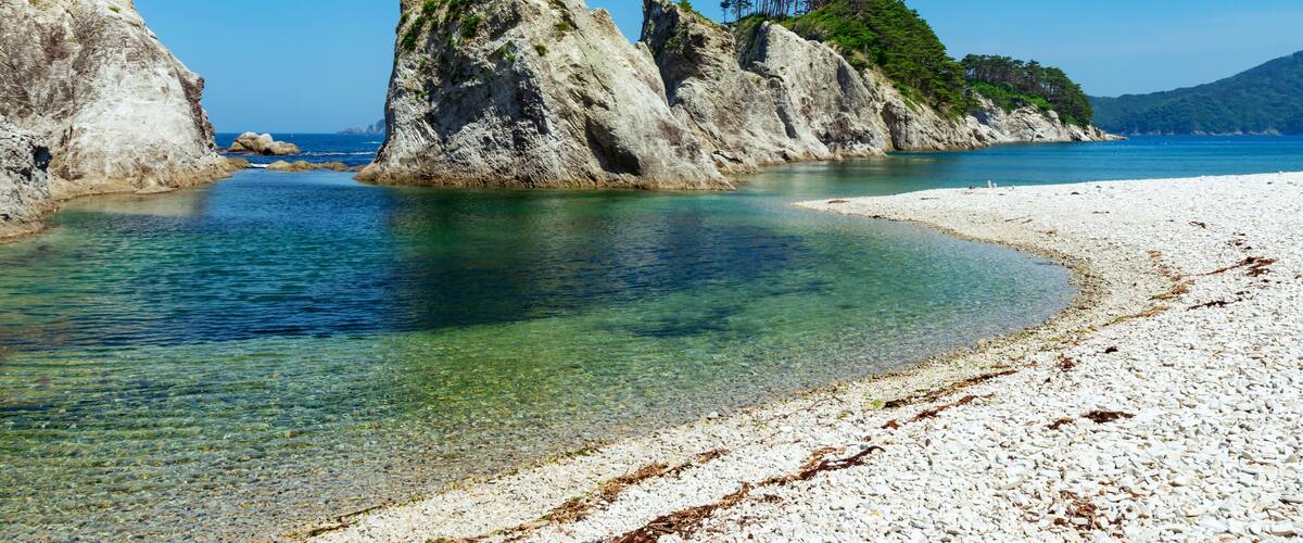 A clear coastal beach with white pebbles, a calm sea, and rocky cliffs with greenery.