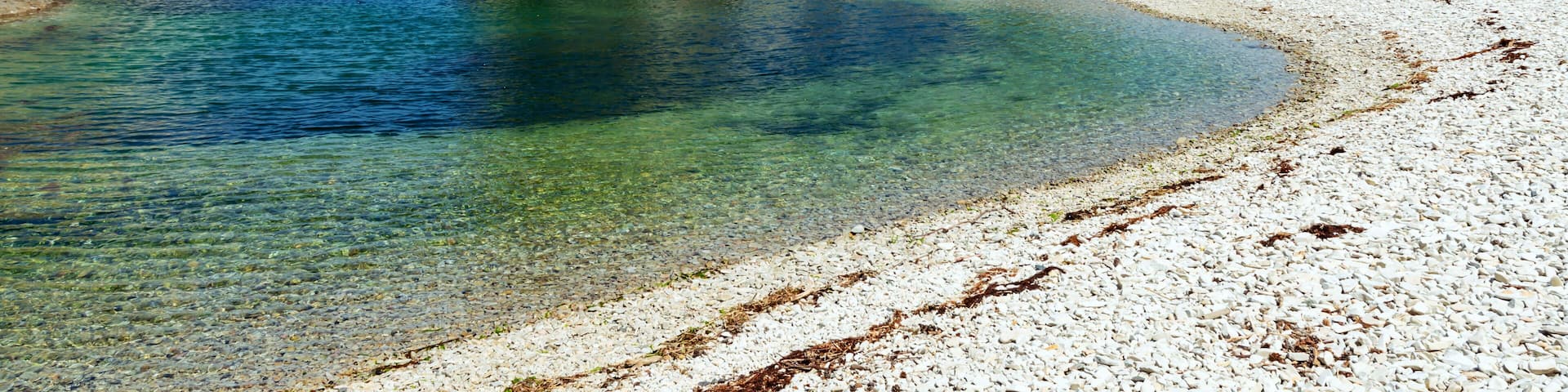 A clear coastal beach with white pebbles, a calm sea, and rocky cliffs with greenery.