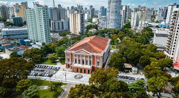 Paisagem urbana com um edifício de telhado vermelho em destaque, rodeado por vegetação e edifícios modernos altos.