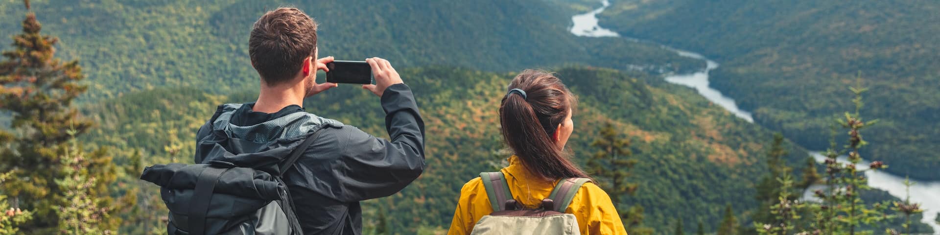 Hikers couple camping walking with backpacks in Quebec taking picture of view with phone in Autumn. Canada forest travel lifestyle banner. Tourists looking at Jacques Cartier National Park.