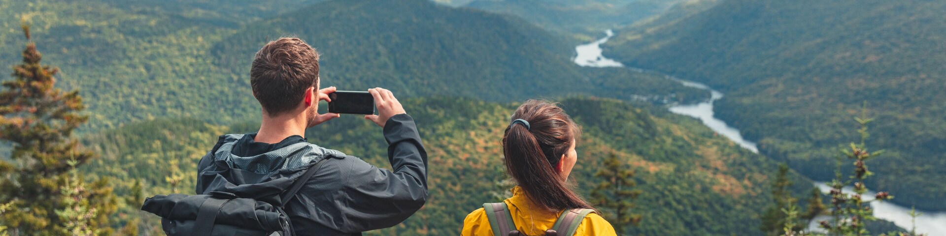 Hikers couple camping walking with backpacks in Quebec taking picture of view with phone in Autumn. Canada forest travel lifestyle banner. Tourists looking at Jacques Cartier National Park.