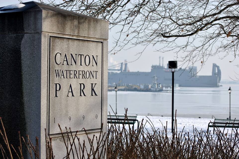 A sign for Canton Waterfront Park with a view of a river and ships in the background.