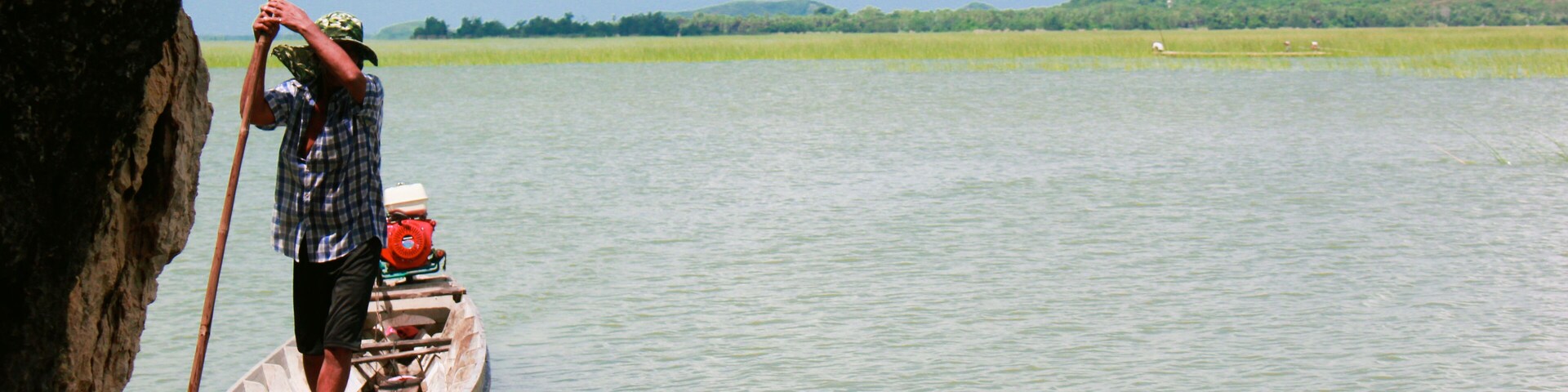 Un hombre en un bote en un río tranquilo con una orilla rocosa y colinas verdes al fondo.
