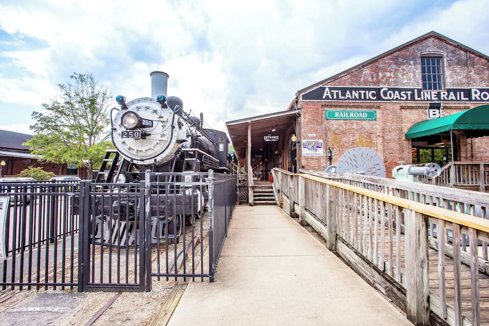 Uma estação de trem histórica com uma locomotiva a vapor imponente, um edifício de tijolos com a inscrição "Atlantic Coast Line Rail Road" e uma área de espera coberta.