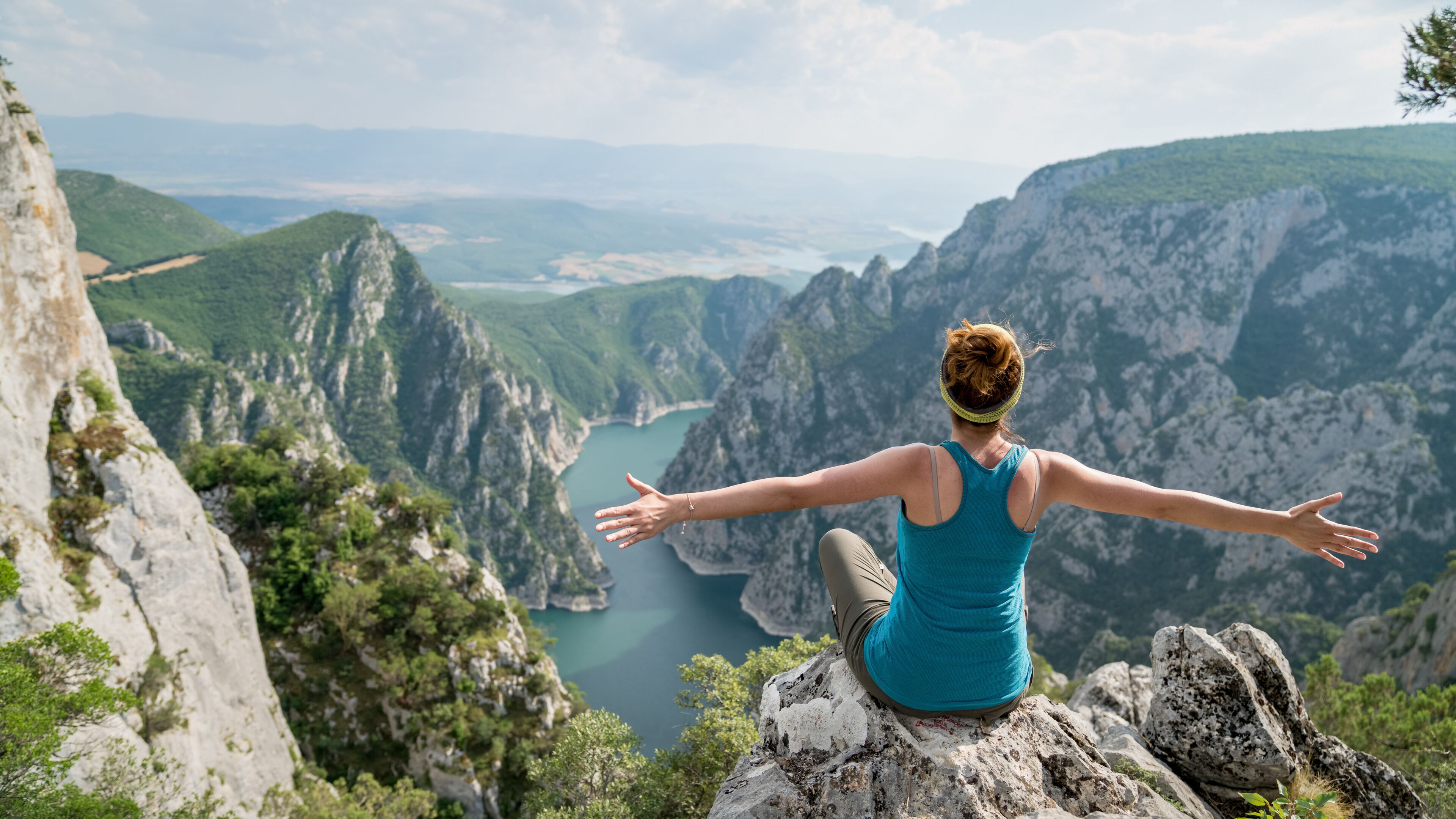 A person sitting on a rock with arms outstretched, overlooking a mountainous landscape with a river.