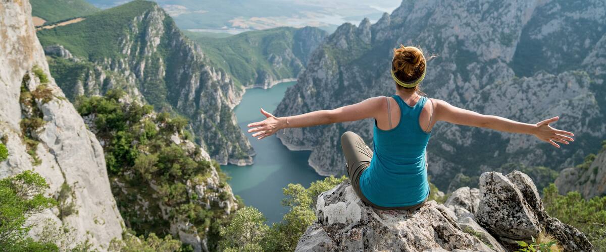 A person sitting on a rock with arms outstretched, overlooking a mountainous landscape with a river.