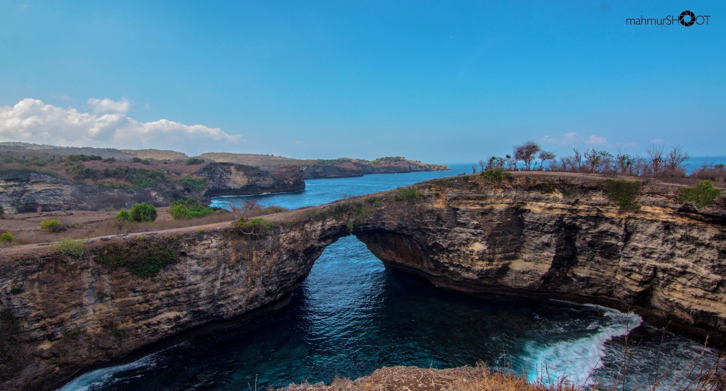 A natural rock arch over a body of water, surrounded by hills and sparse vegetation.