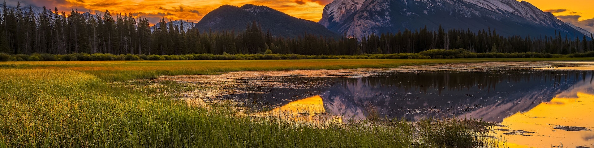 Une chaîne de montagnes au coucher du soleil, avec un lac aux eaux calmes et une zone boisée.