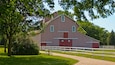 Ein rotes Bauernhaus mit weißen Verzierungen, umgeben von Bäumen und einem weißen Zaun.
