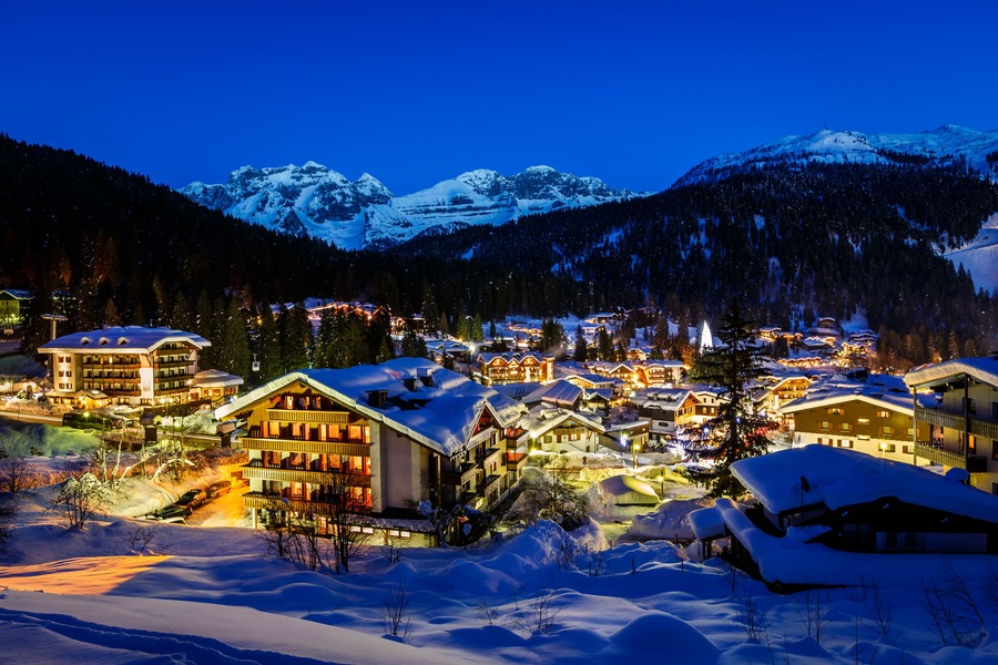 Een besneeuwde bergstad 's nachts, met verlichte gebouwen en een skigebied.
