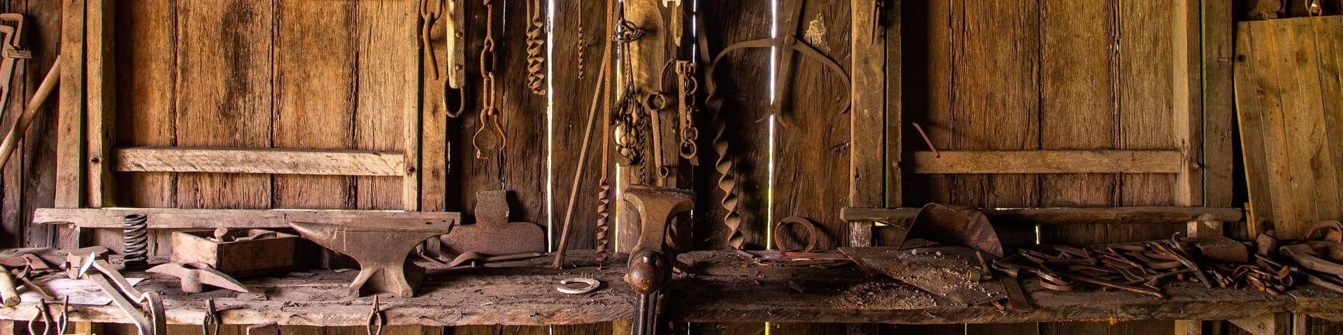 Une vieille forge en bois, avec des outils accrochés aux murs et un établi encombré d’objets métalliques.