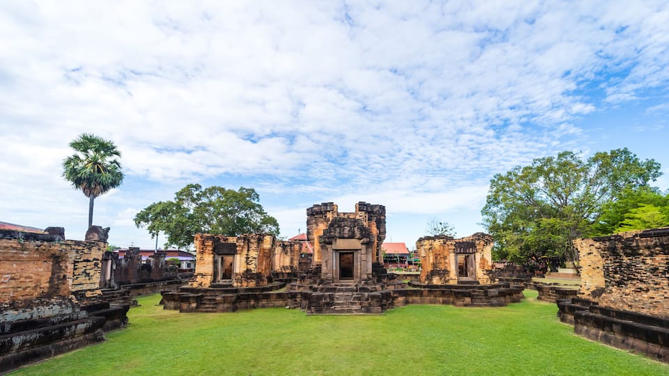 An ancient temple complex with a central entrance, surrounded by stone walls and steps, under a partly cloudy sky.