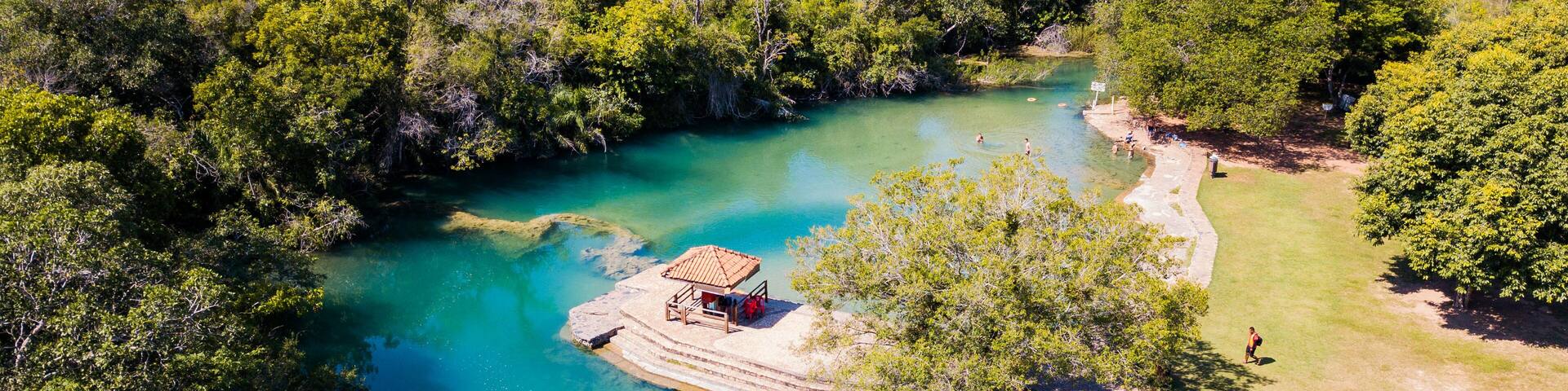 A turquoise river surrounded by dense green forest, a wooden structure on a small island, and a clear path along the riverbank.