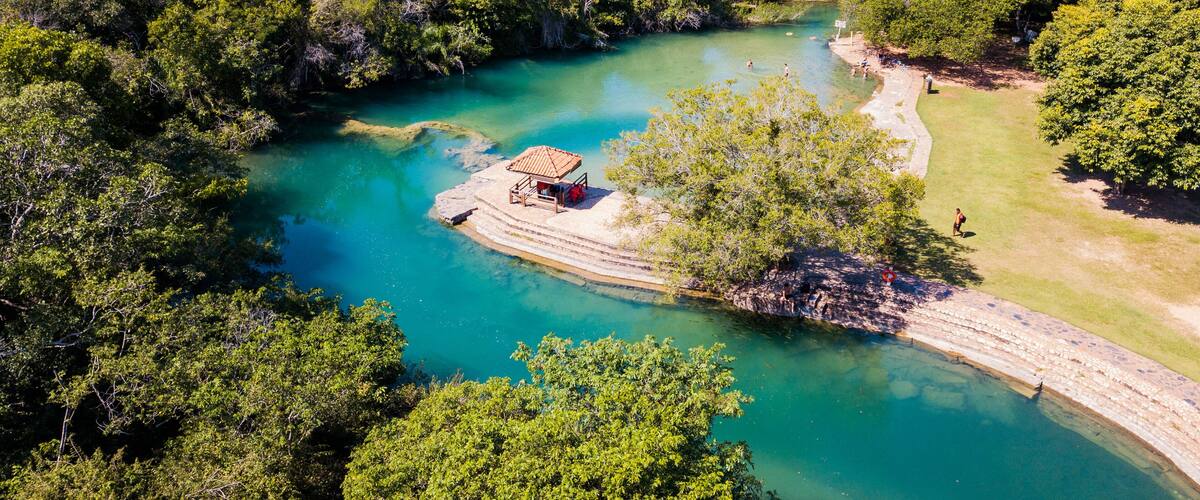 A turquoise river surrounded by dense green forest, a wooden structure on a small island, and a clear path along the riverbank.