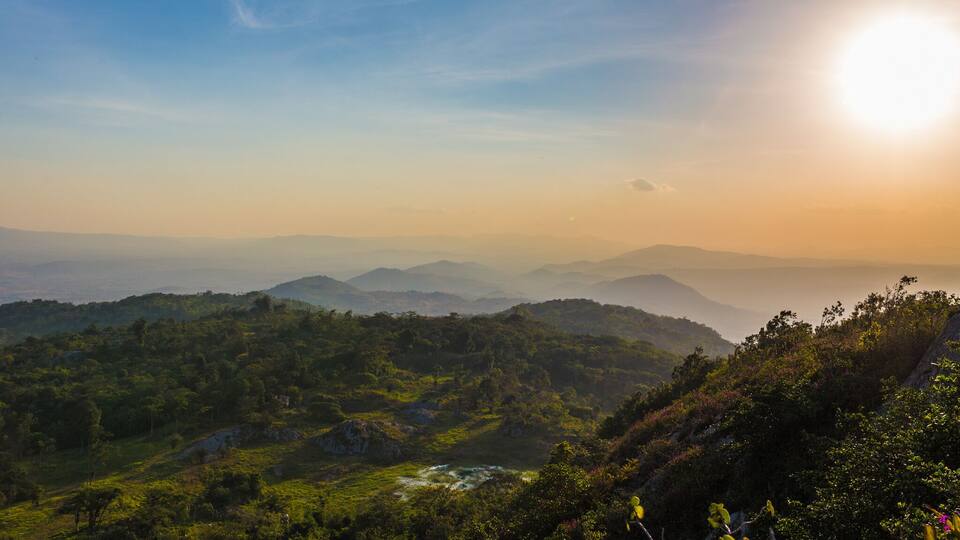 A mountainous landscape with a river, dense greenery, and distant hills under a clear sky.
