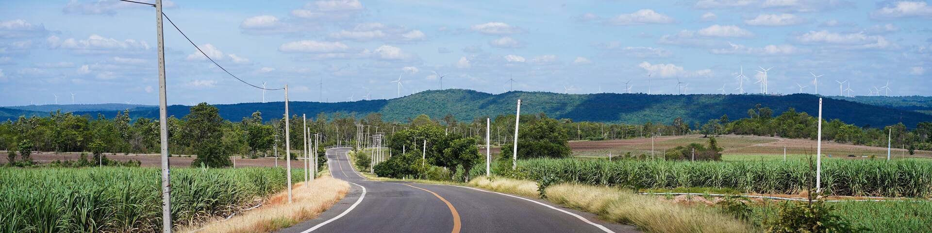 View of concrete road with electric poles in rural lead to windmill turbine on mountain for electric production with blue sky and trees at Chaiyaphum, Thailand. Eco power and energy concepts.