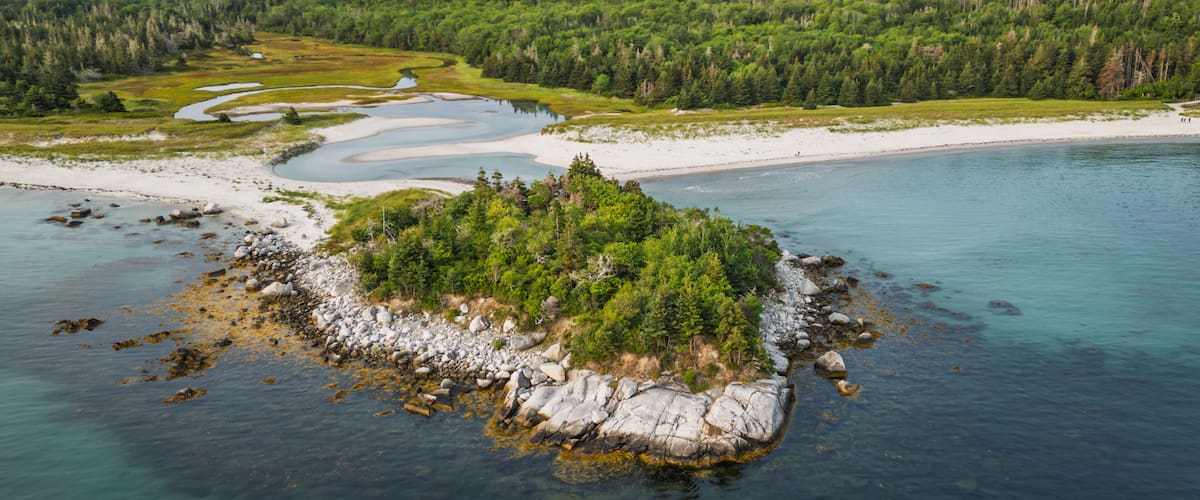 A coastal landscape with a small island surrounded by clear waters, a sandy beach, and dense forest in the background.