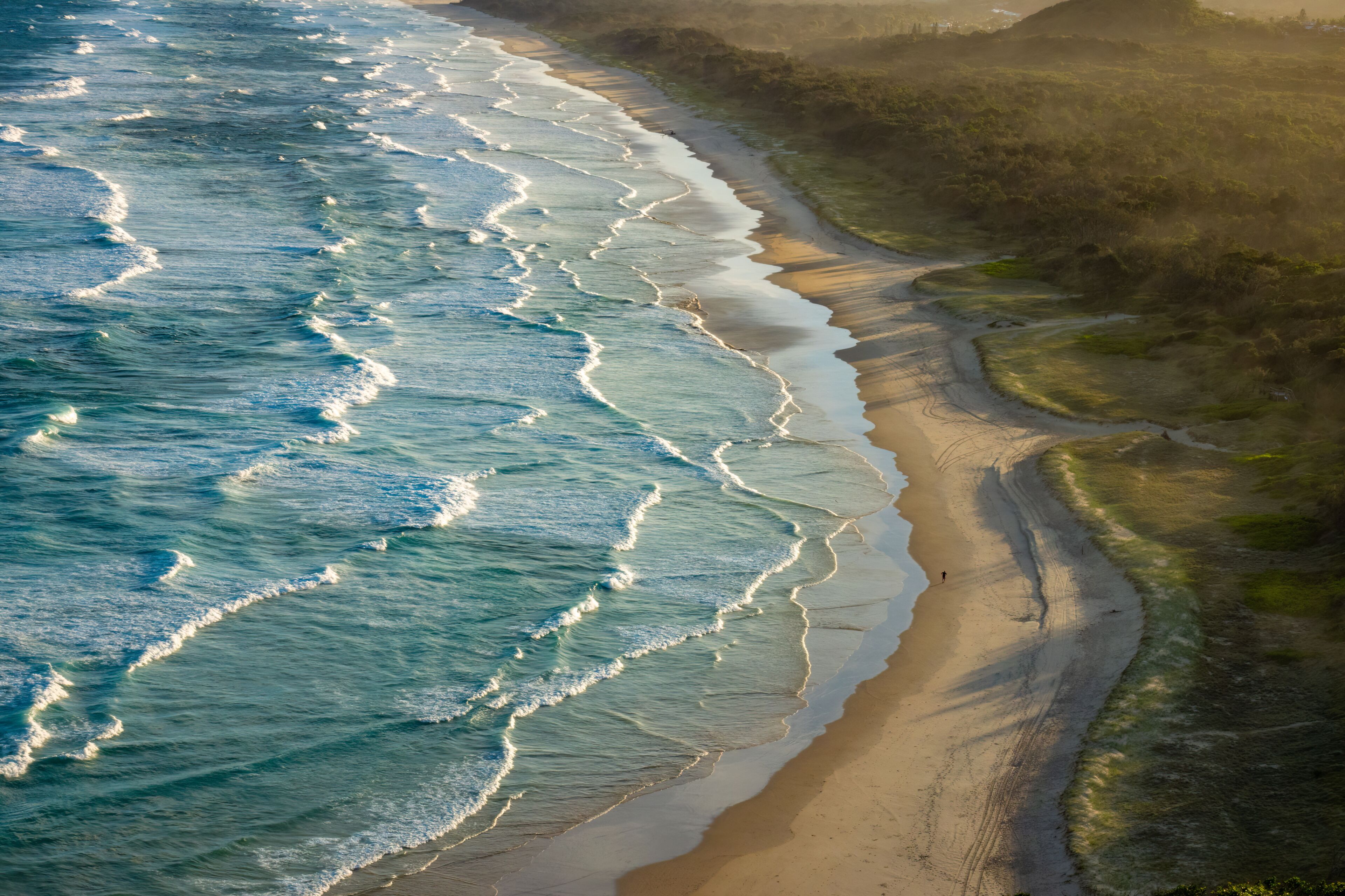 A coastal landscape with waves crashing onto the shore, a sandy beach, and grassy dunes.
