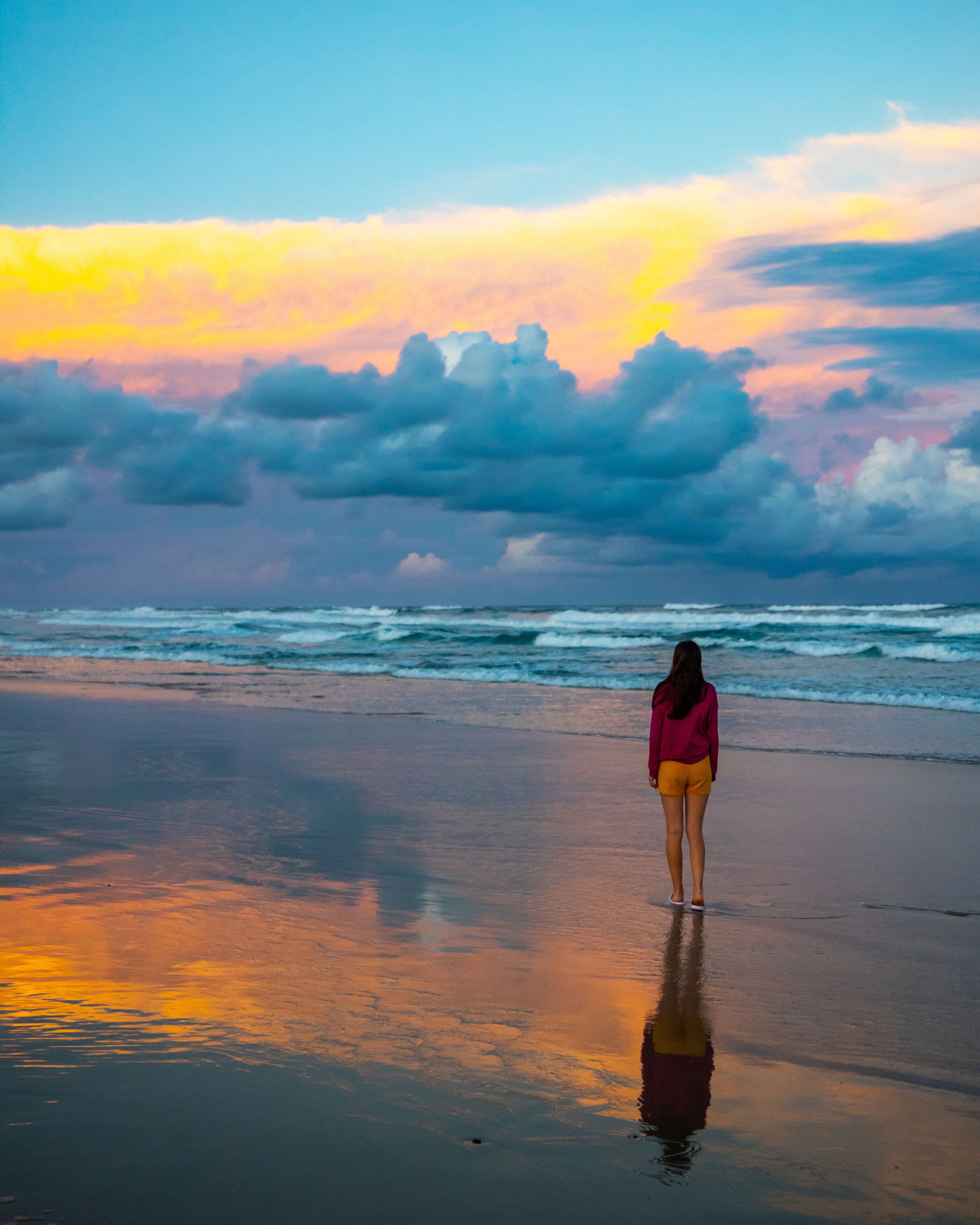 Una playa con una persona de pie mirando al océano, olas rompiendo en la orilla y un cielo de atardecer con nubes.