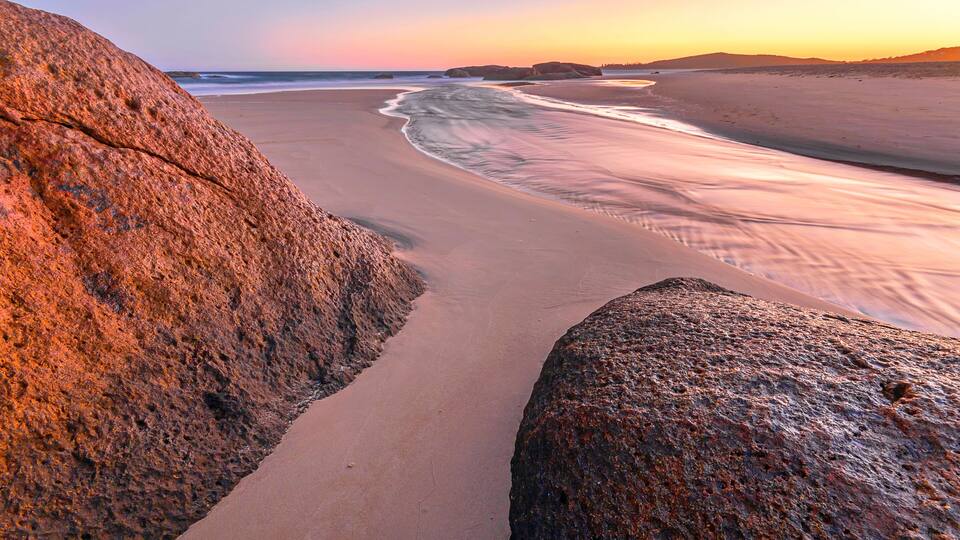 A beach with large rocks and a calm sea at sunset.