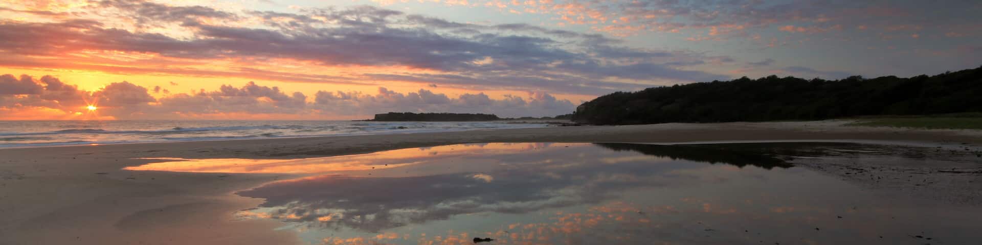 A beach with a sunset, calm waters, and a distant landmass.