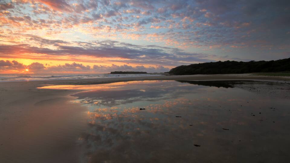 A beach with a sunset, calm waters, and a distant landmass.