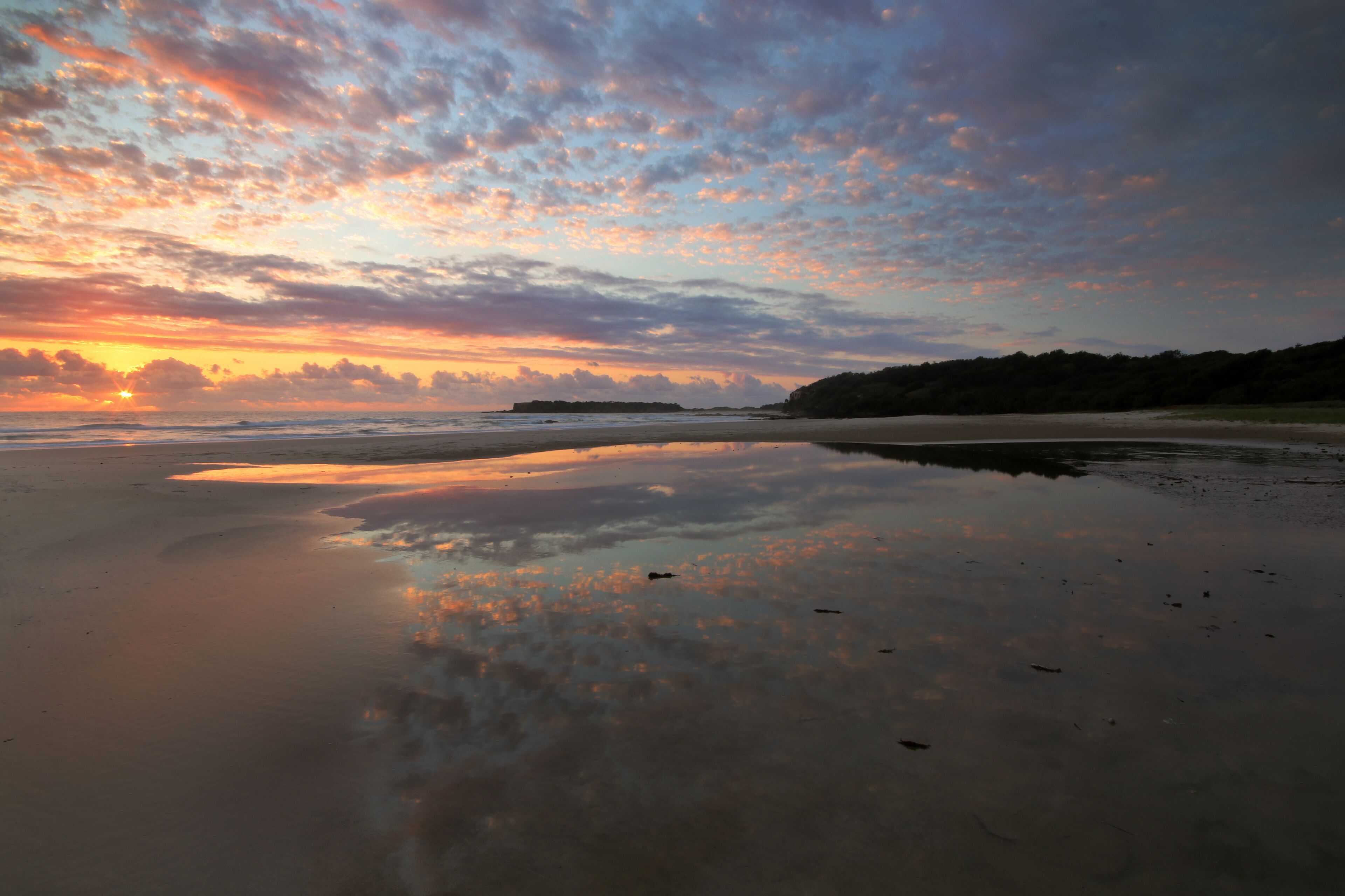 A beach with a sunset, calm waters, and a distant landmass.
