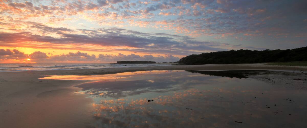 A beach with a sunset, calm waters, and a distant landmass.