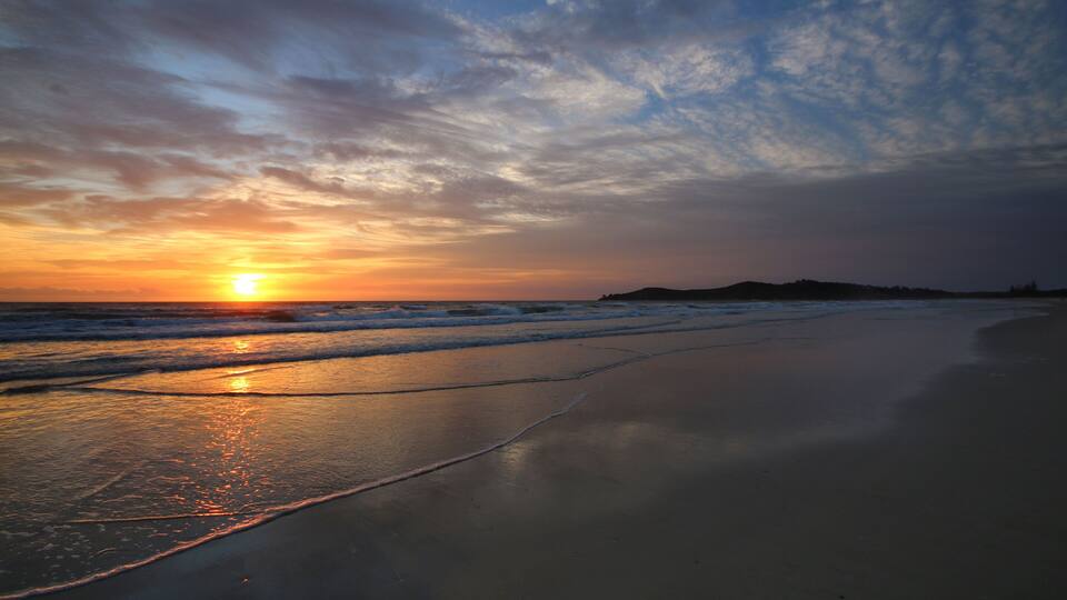 A sunset over a beach with waves, a clear sky, and distant hills.