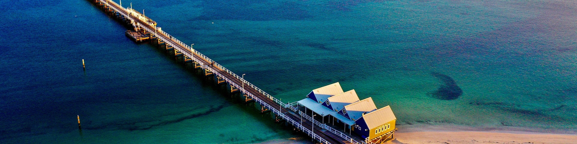 Aerial view of Busselton Jetty extending into the turquoise waters of Geographe Bay at sunset, with wooden planks and a small building visible in Busselton, Western Australia, Australia.