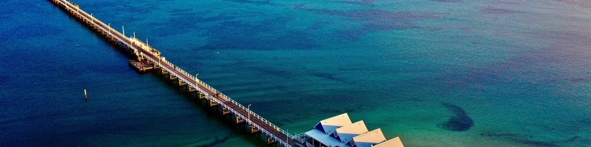 Aerial view of Busselton Jetty extending into the turquoise waters of Geographe Bay at sunset, with wooden planks and a small building visible in Busselton, Western Australia, Australia.
