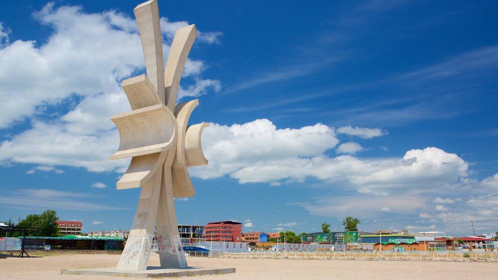 Eine große, abstrakte Skulptur in einer sandigen Umgebung, im Hintergrund sind Gebäude zu sehen.