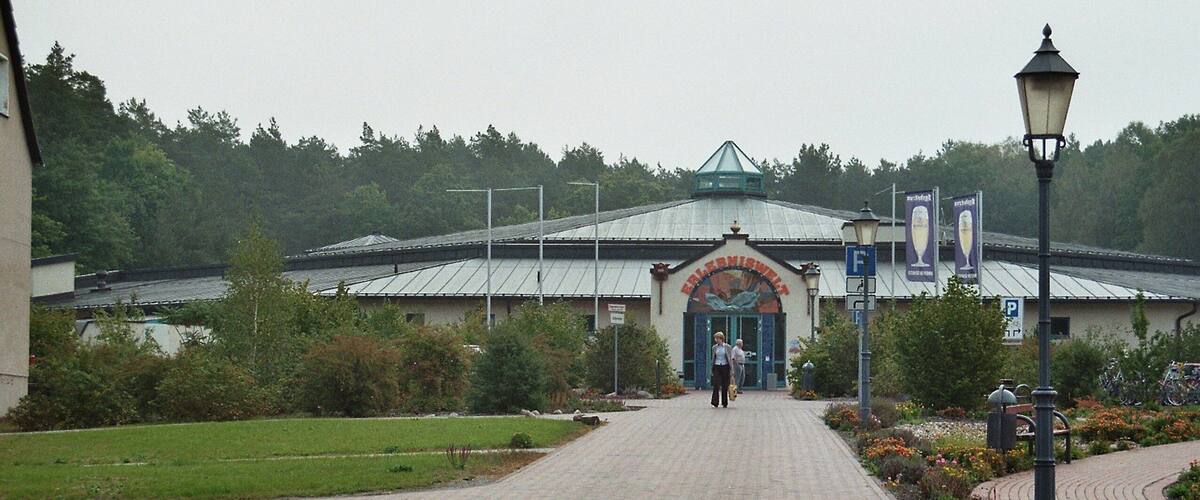 A paved pathway leading to a building with a green roof and an arched entrance. There are street lamps and a sign with a 'P' parking symbol.