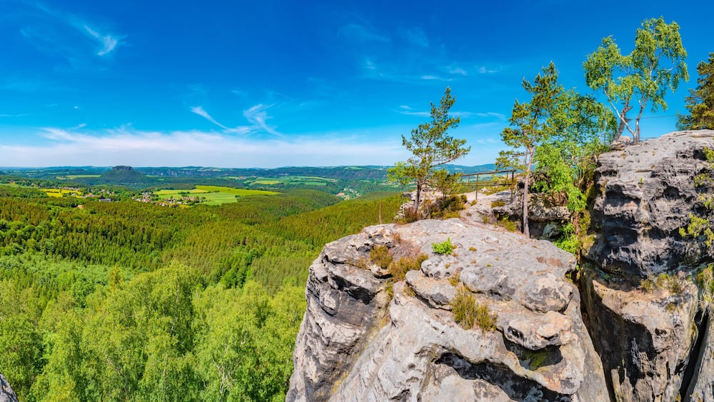 Eine gebirgige Landschaft mit einem bewaldeten Tal, blauem Himmel und einem felsigen Vorsprung im Vordergrund.