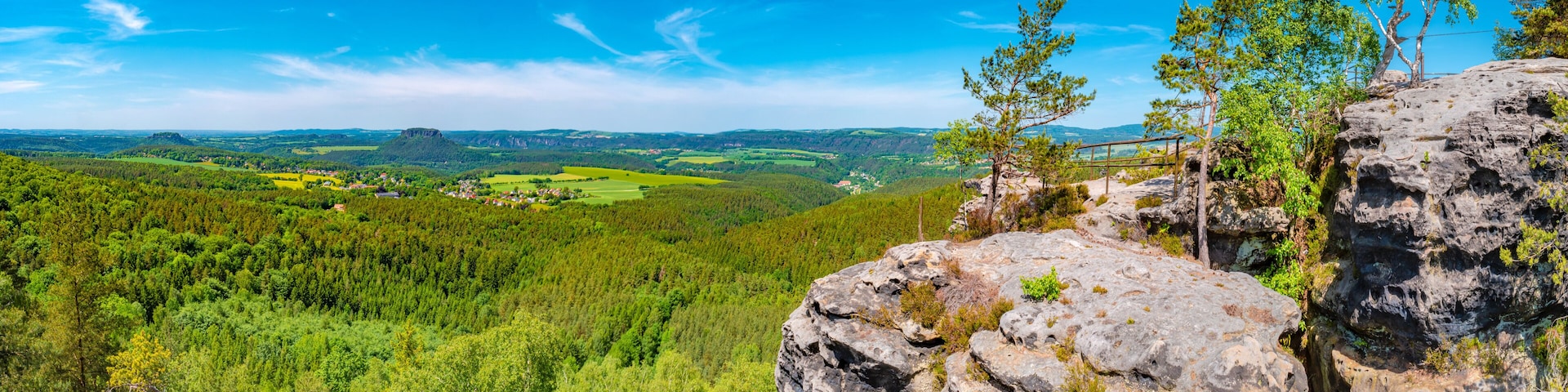 Paisagem montanhosa com um vale arborizado, céu azul límpido e um afloramento rochoso em primeiro plano.