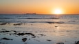 Ein Sonnenuntergang über einem Strand mit verstreuten Felsen und Gebäuden in der Ferne.