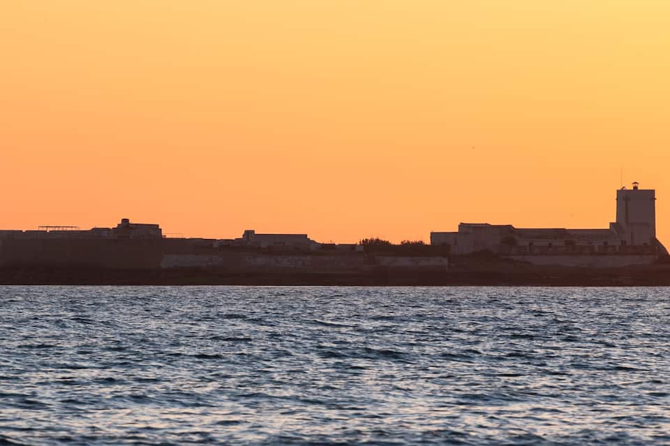 A coastal fortification with a prominent tower, set against a sunset sky.