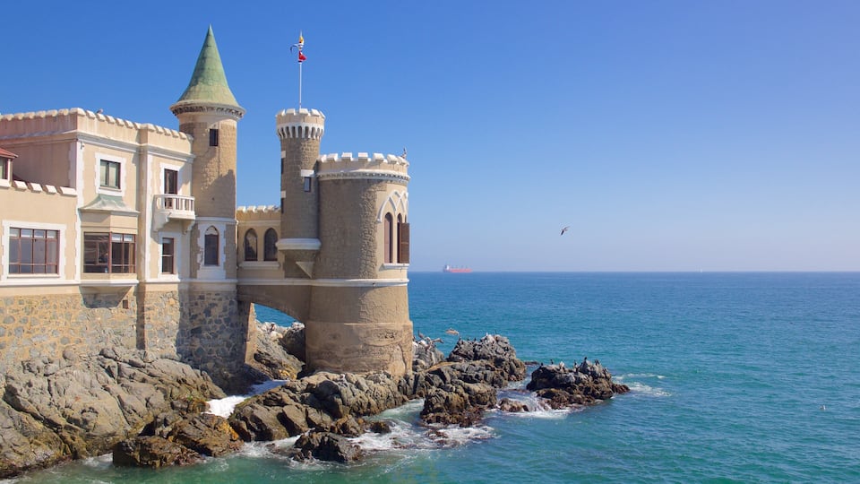 A coastal castle with a green spire and a red flag, overlooking the sea with a ship in the distance.