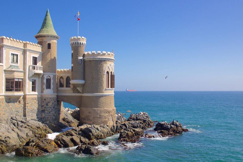 Un castillo costero con un pináculo verde y una bandera roja, que domina el mar con un barco a lo lejos.